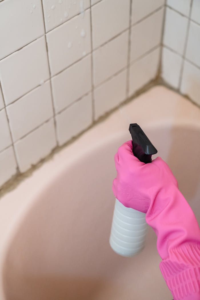 A person wearing a pink glove cleans bathroom tiles with a spray bottle.